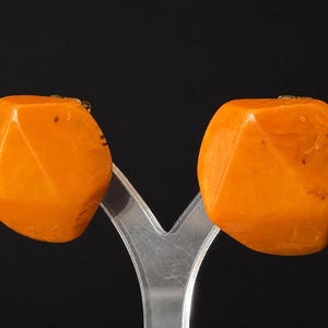 May include: A pair of clip-on earrings featuring large, faceted, orange resin stones with a marbled texture. The stones have a geometric, multi-sided shape with smooth, polished surfaces and visible internal variations in the orange color.