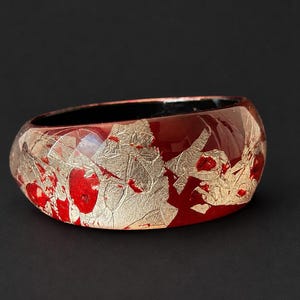 May include: A wide bangle bracelet featuring a glossy deep red base with irregular, reflective silver-toned metallic leaf flakes embedded across the outer surface and a smooth, solid black interior lining.