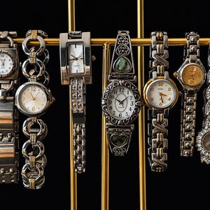 May include: A selection of ladies' watches displayed on a gold-coloured stand against a black backdrop. The watches showcase diverse designs, including silver and gold tones, with round and rectangular faces. Some watches have decorative embellishments.