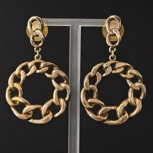 May include: A pair of gold-toned chain link earrings. Each earring features a circular chain design suspended from a smaller chain link and a post. The earrings are displayed against a dark background.