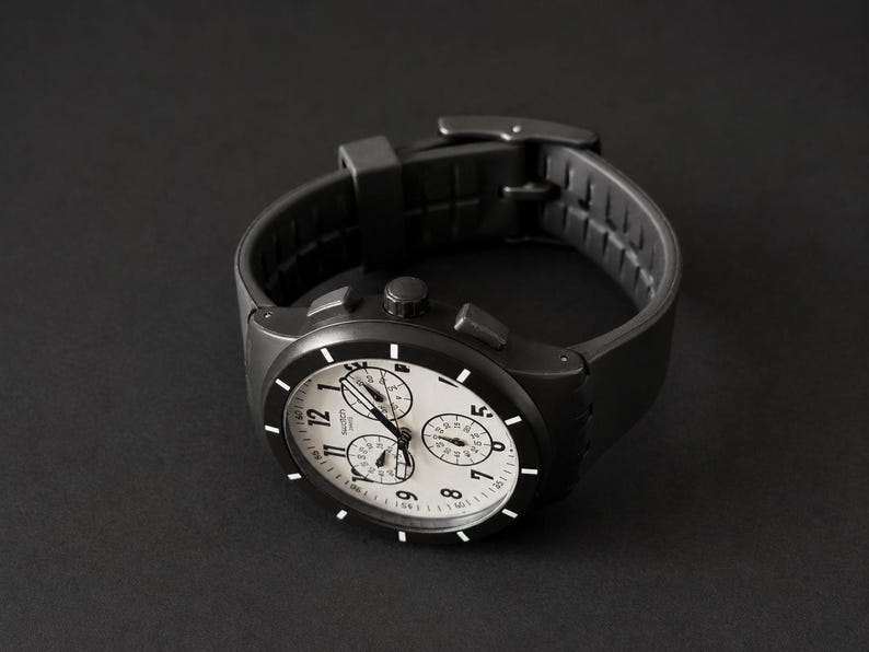 K&ouml;nnte beinhalten: Eine schwarze Armbanduhr mit wei&szlig;em Zifferblatt und schwarzen Zeigern. Die Uhr hat drei kleinere Zifferbl&auml;tter und ein schwarzes Kautschukarmband. Die Uhr ist vor einem dunklen Hintergrund platziert.