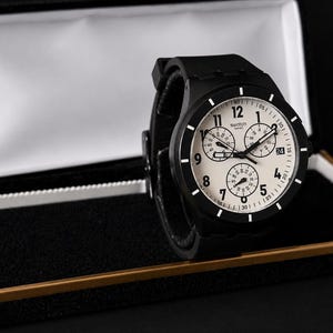 K&ouml;nnte beinhalten: Eine schwarze Swatch-Uhr mit wei&szlig;em Zifferblatt und drei Hilfszifferbl&auml;ttern, pr&auml;sentiert in einer offenen schwarzen und goldenen Uhrenbox. Die Uhr hat ein schwarzes Armband und ein rundes Zifferblatt mit wei&szlig;en Ziffern und Markierungen. Ein stilvolles Accessoire.