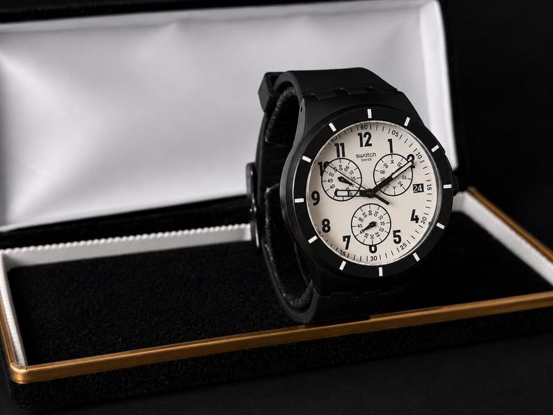 K&ouml;nnte beinhalten: Eine schwarze Swatch-Uhr mit wei&szlig;em Zifferblatt und drei Hilfszifferbl&auml;ttern, pr&auml;sentiert in einer offenen schwarzen und goldenen Uhrenbox. Die Uhr hat ein schwarzes Armband und ein rundes Zifferblatt mit wei&szlig;en Ziffern und Markierungen. Ein stilvolles Accessoire.