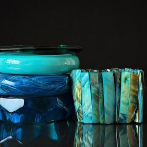 May include: Four bracelets in varying shades of blue and teal are stacked. The bracelets include a dark teal bangle, a bright turquoise bangle, a marbled blue bangle, and a faceted dark blue bangle. A separate bracelet is made of rectangular teal and gold pieces.