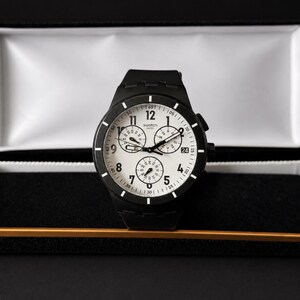 K&ouml;nnte beinhalten: Eine schwarze Armbanduhr mit wei&szlig;em Zifferblatt und drei Hilfszifferbl&auml;ttern, pr&auml;sentiert in einer offenen schwarzen und goldenen Uhrenbox. Die Uhr hat ein schwarzes Armband und wei&szlig;e Ziffern. Das Zifferblatt zeigt die Zahlen 1 bis 12.