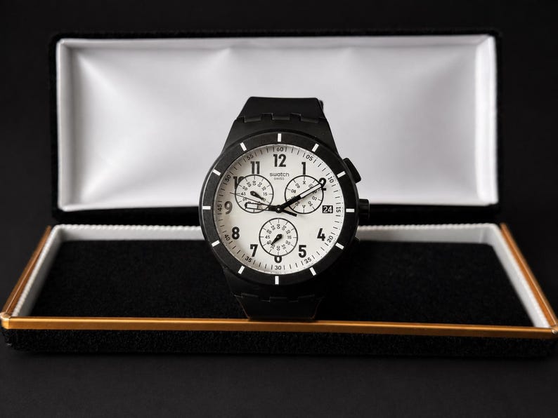 K&ouml;nnte beinhalten: Eine schwarze Armbanduhr mit wei&szlig;em Zifferblatt und drei Hilfszifferbl&auml;ttern, pr&auml;sentiert in einer offenen schwarzen und goldenen Uhrenbox. Die Uhr hat ein schwarzes Armband und wei&szlig;e Ziffern. Das Zifferblatt zeigt die Zahlen 1 bis 12.