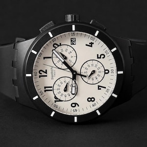 K&ouml;nnte beinhalten: Eine schwarze Swatch Chronographenuhr mit cremefarbenem Zifferblatt. Die Uhr verf&uuml;gt &uuml;ber drei Hilfszifferbl&auml;tter, ein Datumsfenster und wei&szlig;e Stundenmarkierungen. Die Uhr hat eine schwarze L&uuml;nette und ein schwarzes Kautschukarmband.