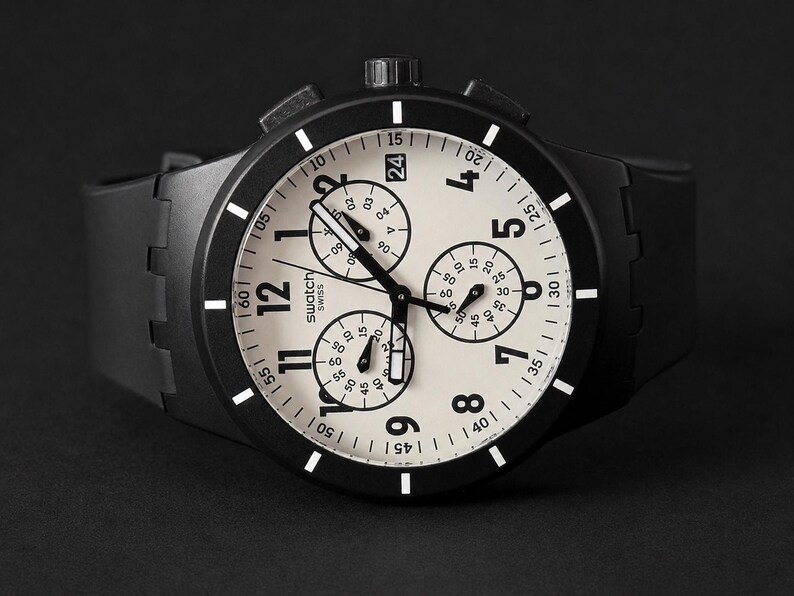K&ouml;nnte beinhalten: Eine schwarze Swatch Chronographenuhr mit cremefarbenem Zifferblatt. Die Uhr verf&uuml;gt &uuml;ber drei Hilfszifferbl&auml;tter, ein Datumsfenster und wei&szlig;e Stundenmarkierungen. Die Uhr hat eine schwarze L&uuml;nette und ein schwarzes Kautschukarmband.