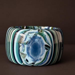 Könnte beinhalten: Ein breiter Armreif mit einem wirbelnden Muster aus tiefblauem, waldgrünem, blaugrünem und weißem Glas. Die Oberfläche zeigt einen markanten, texturierten Mittelpunkt mit kraterartigem Aussehen, umgeben von glatten, vertikalen Farbbändern.