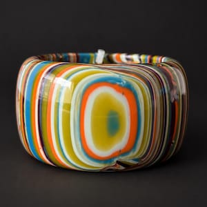 May include: A wide, cylindrical bangle bracelet crafted from layered, multi-colored glass. The surface features concentric, swirling bands of vibrant orange, lime green, bright blue, white, and deep purple, creating a smooth, polished finish.