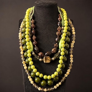 May include: A multi-strand statement necklace featuring a variety of beads in shades of lime green, olive green, brown, and clear crystal. The design incorporates round, faceted, and textured beads, including a central square-shaped brown bead.