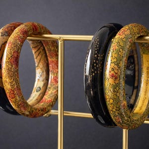 May include: Four decorative bangles on a gold-coloured display stand. The bangles feature floral and abstract patterns in shades of black, yellow, orange, and red. The stand has a simple, elegant design.