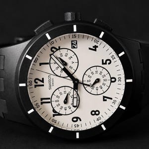 K&ouml;nnte beinhalten: Schwarze Swatch Chronographenuhr mit wei&szlig;em Zifferblatt und schwarzen Akzenten. Die Uhr verf&uuml;gt &uuml;ber drei Hilfszifferbl&auml;tter, eine Datumsanzeige und ein schwarzes Kautschukarmband. Das Zifferblatt zeigt das Swatch Swiss Logo.