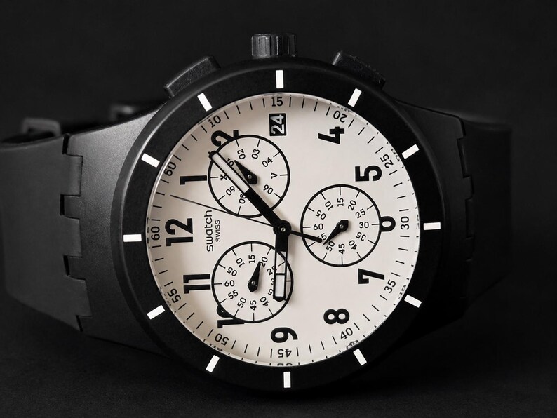 K&ouml;nnte beinhalten: Schwarze Swatch Chronographenuhr mit wei&szlig;em Zifferblatt und schwarzen Akzenten. Die Uhr verf&uuml;gt &uuml;ber drei Hilfszifferbl&auml;tter, eine Datumsanzeige und ein schwarzes Kautschukarmband. Das Zifferblatt zeigt das Swatch Swiss Logo.