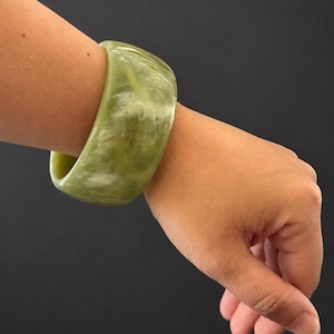 May include: A wide, polished bangle bracelet featuring a marbled pattern of sage green and creamy white tones. The bracelet has a smooth, rounded exterior surface and a thick, solid construction, displaying a glossy finish throughout the piece.