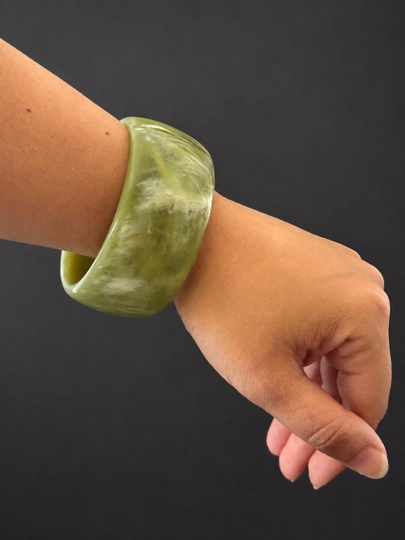 May include: A wide, polished bangle bracelet featuring a marbled pattern of sage green and creamy white tones. The bracelet has a smooth, rounded exterior surface and a thick, solid construction, displaying a glossy finish throughout the piece.