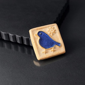 Peut inclure: Pendentif carré en céramique avec un motif d'oiseau bleu. L'oiseau est centré sur un fond beige, avec un petit soleil dans le coin supérieur droit. Le pendentif mesure environ 4 cm de côté.