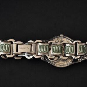 May include: A silver-toned Swatch watch with a clear face and a matching metal band. The band features rectangular links with green enamel detailing. The watch is set against a black background.