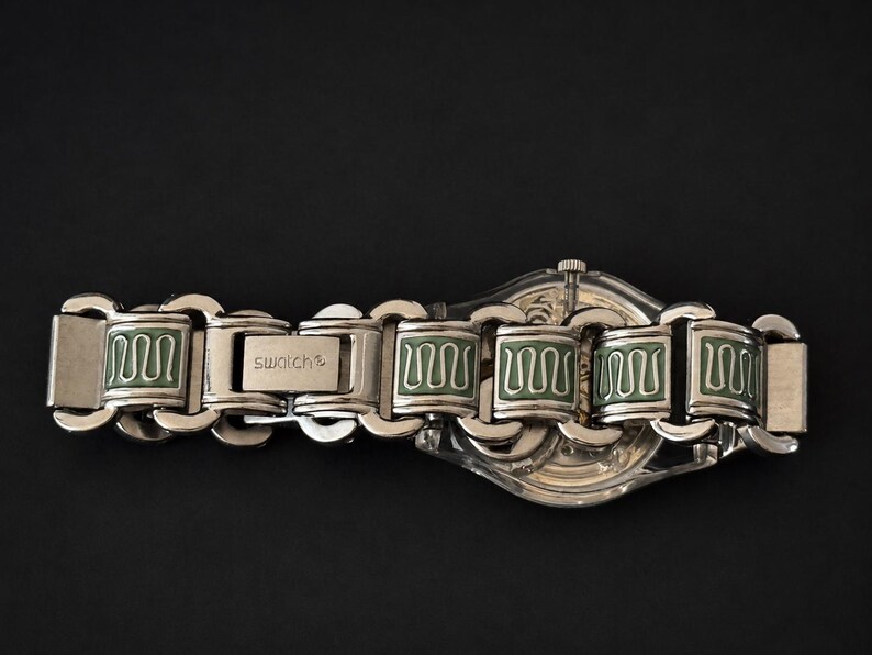 May include: A silver-toned Swatch watch with a clear face and a matching metal band. The band features rectangular links with green enamel detailing. The watch is set against a black background.