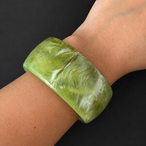 May include: A wide bangle bracelet featuring a polished, translucent lime green surface with swirling white marbled patterns throughout the material. The bracelet has a smooth, rounded rectangular profile and a glossy finish that reflects light.