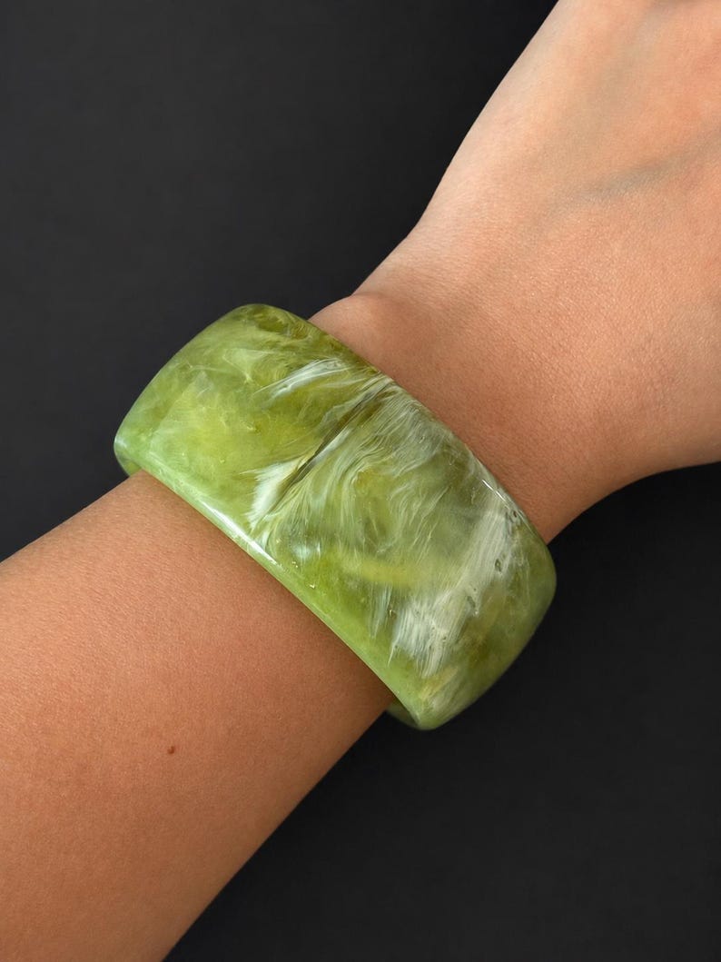 May include: A wide bangle bracelet featuring a polished, translucent lime green surface with swirling white marbled patterns throughout the material. The bracelet has a smooth, rounded rectangular profile and a glossy finish that reflects light.