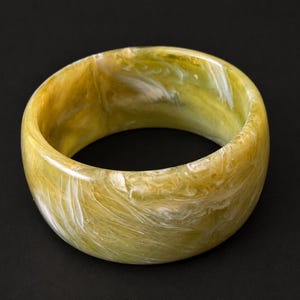 May include: A wide, circular bangle bracelet featuring a polished, marbled finish with swirling patterns of olive green, mustard yellow, and creamy white tones throughout the smooth, rounded resin surface of the jewelry piece.
