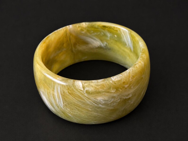 May include: A wide, circular bangle bracelet featuring a polished, marbled finish with swirling patterns of olive green, mustard yellow, and creamy white tones throughout the smooth, rounded resin surface of the jewelry piece.