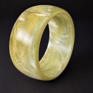 May include: A wide, circular bangle bracelet featuring a translucent, pale yellow and cream-colored marbled pattern. The surface of the bracelet is smooth and polished, displaying swirling, cloud-like textures throughout the material.
