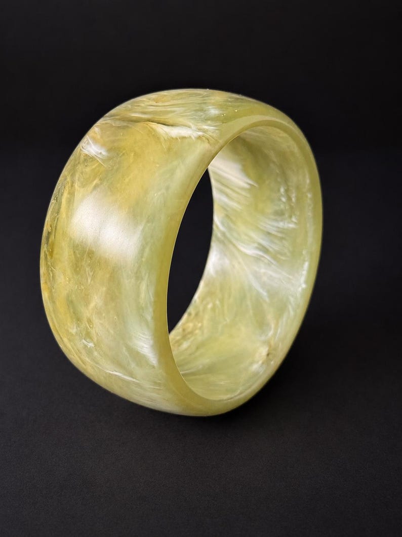 May include: A wide, circular bangle bracelet featuring a translucent, pale yellow and cream-colored marbled pattern. The surface of the bracelet is smooth and polished, displaying swirling, cloud-like textures throughout the material.