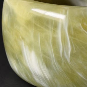 May include: A wide bangle bracelet featuring a smooth, polished surface with a marbled pattern of pale olive green and creamy white swirls. The material has a translucent quality, creating depth and light reflection across the curved exterior.