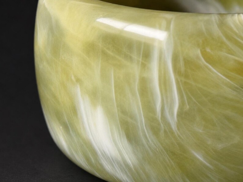 May include: A wide bangle bracelet featuring a smooth, polished surface with a marbled pattern of pale olive green and creamy white swirls. The material has a translucent quality, creating depth and light reflection across the curved exterior.