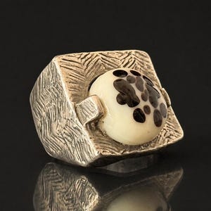 May include: A silver ring with a textured square setting. The ring features a cream-coloured, rounded stone with dark brown spots. The ring is set against a black background, with a reflection visible below.