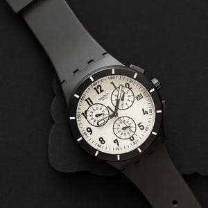 K&ouml;nnte beinhalten: Eine schwarze Swatch Chronographenuhr mit wei&szlig;em Zifferblatt und schwarzen Zeigern. Die Uhr verf&uuml;gt &uuml;ber mehrere Hilfszifferbl&auml;tter und ein schwarzes Kautschukarmband. Das Zifferblatt zeigt das Swatch-Logo und verschiedene Zahlen.