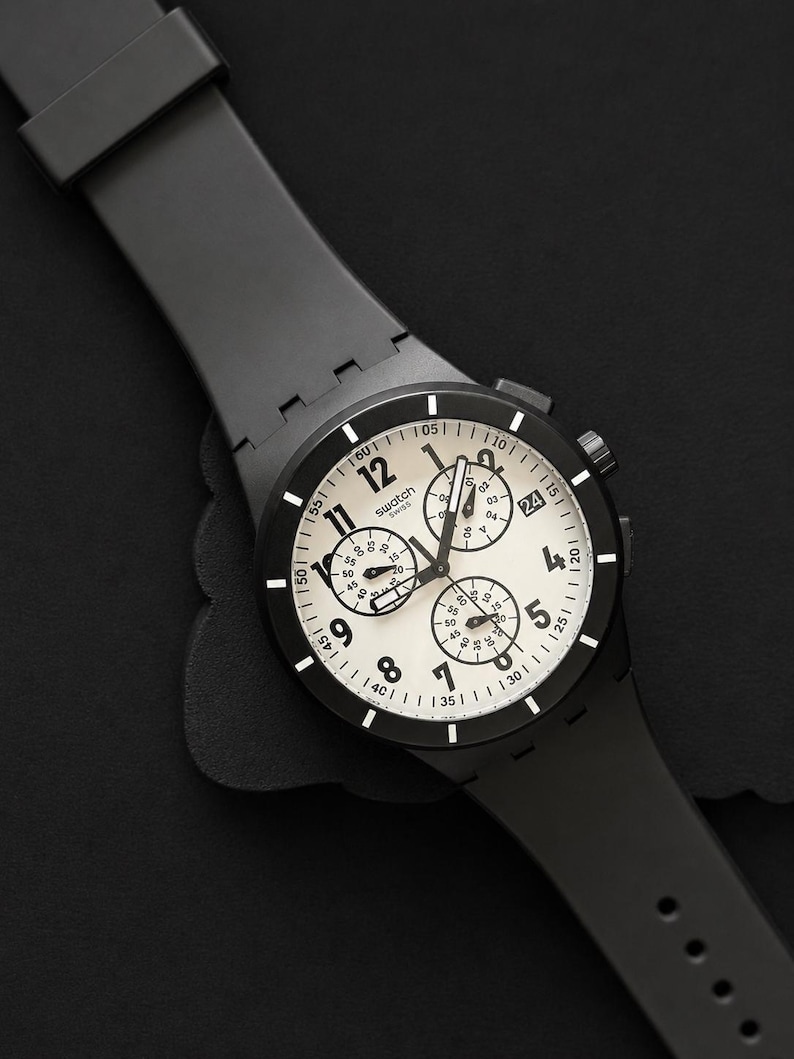 K&ouml;nnte beinhalten: Eine schwarze Swatch Chronographenuhr mit wei&szlig;em Zifferblatt und schwarzen Zeigern. Die Uhr verf&uuml;gt &uuml;ber mehrere Hilfszifferbl&auml;tter und ein schwarzes Kautschukarmband. Das Zifferblatt zeigt das Swatch-Logo und verschiedene Zahlen.