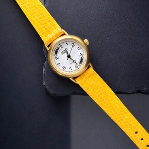 May include: A gold-toned wristwatch with a white face and black hands, featuring the brand name "GITANE". The watch has a bright yellow textured leather band. The watch is set against a dark background.