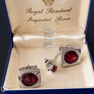 May include: A set of silver-toned square cufflinks and a matching tie tack, each featuring a faceted deep red stone. The items are presented in a blue hinged box with the text Royal Standard Imported Stones printed on the interior lid.