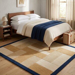 May include: A rectangular area rug featuring a geometric pattern of tan, beige, and cream-coloured blocks, framed by a solid dark blue border. The rug is positioned on a light-coloured wooden floor beneath a wooden bed frame with bedding.