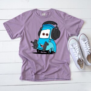 Op de afbeelding: Een lila T-shirt met een cartoonafbeelding van een blauw autokarakter met zwarte oorbeschermers. De auto heeft een lachend gezicht en houdt een gereedschap vast. Witte sneakers zijn rechts van het shirt geplaatst.