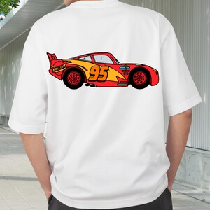 Op de afbeelding: Wit T-shirt met een kleurrijke cartoon illustratie van een rood-gele raceauto met nummer 95. Het auto-ontwerp bevat het woord "Rusteze" en zwarte wielen. Dit is een casual kledingstuk.