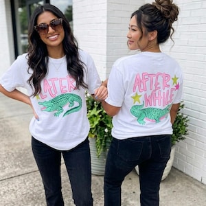 Peut inclure: Deux personnes portant des t-shirts blancs avec un motif d'alligator vert. Le devant du t-shirt porte l'inscription "LATER" en rose. Le dos du t-shirt indique "AFTER WHILE" en lettres roses avec des étoiles.