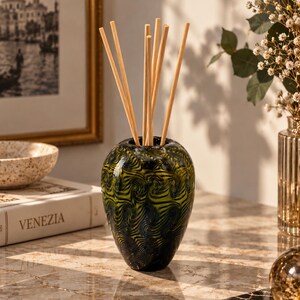 May include: A glass reed diffuser vessel featuring a swirling pattern of dark green and black colors. The rounded, bulbous container holds several light-colored wooden reeds extending upwards from the narrow opening at the top of the glass piece.
