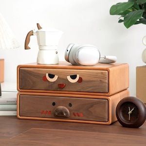 May include: A two-drawer wooden storage unit with cartoon-like faces on the drawers. A white espresso maker, gray headphones, and a small wooden clock sit on top. The drawers are brown with light brown frames.