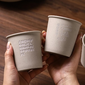 Puede incluir: Dos tazas beige con diseños en relieve. Una taza presenta el alfabeto, y la otra texto y un símbolo triangular. Las tazas tienen un aspecto texturizado y una forma cilíndrica simple. Las tazas se sostienen en las manos.
