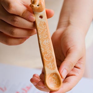 Peut inclure: Une cuillère en céramique marron clair intitulée Doodled Bread Spoon avec des dessins au trait blancs gravés d'étoiles, de cœurs et de cercles. La cuillère mesure environ 13 cm de long et 2,5 cm de large, avec une petite tête d'animal sculptée.
