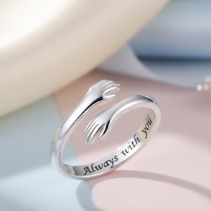 May include: A silver-colored ring with two arms embracing. The ring has the words "Always with you" engraved on the band. The ring is on a pink and blue surface.