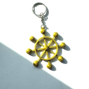Może przedstawiać: Złoty brelok do kluczy z wystylizowanym wzorem koła z ośmioma sferycznymi akcentami. Koło jest połączone ze srebrnym łańcuszkiem i kółkiem na klucze. Brelok znajduje się na białym tle.
