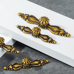 Peut inclure: Quatre poignées de meubles en laiton antique avec un motif floral décoratif. Chaque poignée présente un bouton central et des détails ornés. Les poignées sont disposées sur une surface blanche sur fond gris.