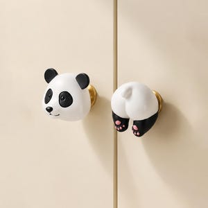 May include: Two panda-themed cabinet knobs. One knob features the panda's face with black ears and eyes, and a white face. The other knob shows the panda's rear, with black legs and pink paw pads. Both knobs have gold-colored bases.