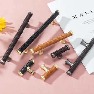 Pode incluir: Uma coleção de puxadores decorativos para armários em várias cores, incluindo castanho, preto e bege. Os puxadores apresentam um design em couro trançado e detalhes metálicos. Alguns são longos, outros mais curtos. A palavra "MALIBU" é visível num papel.