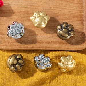 Könnte beinhalten: Sechs pfotenförmige Schrankknöpfe in Silber-, Gold- und Bronze-Ausführungen. Die Knöpfe sind auf einem Holztablett vor gelbem Hintergrund angeordnet. Die Knöpfe sind als Katzenpfoten gestaltet. Geeignet für Möbel.